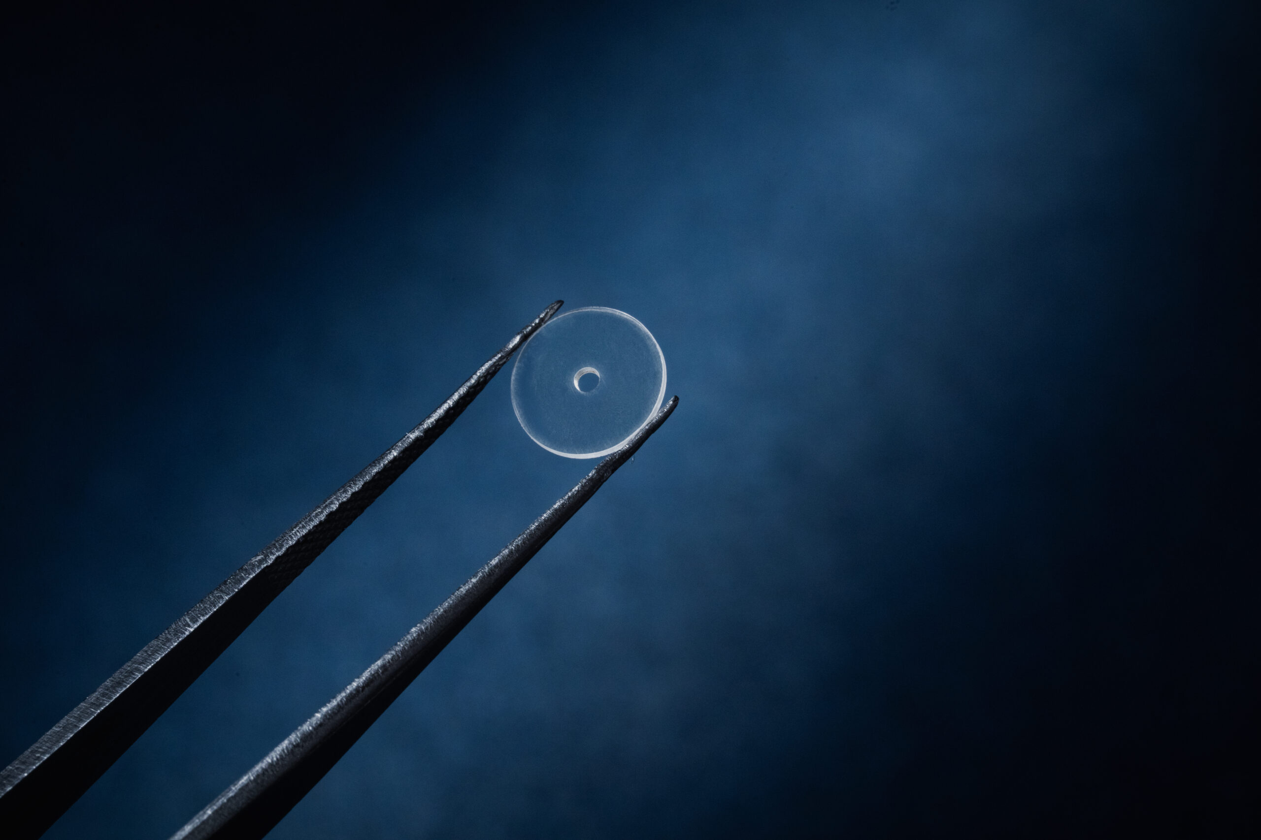 Two tweezers holding a transparent circular disc with central aperture against blue background